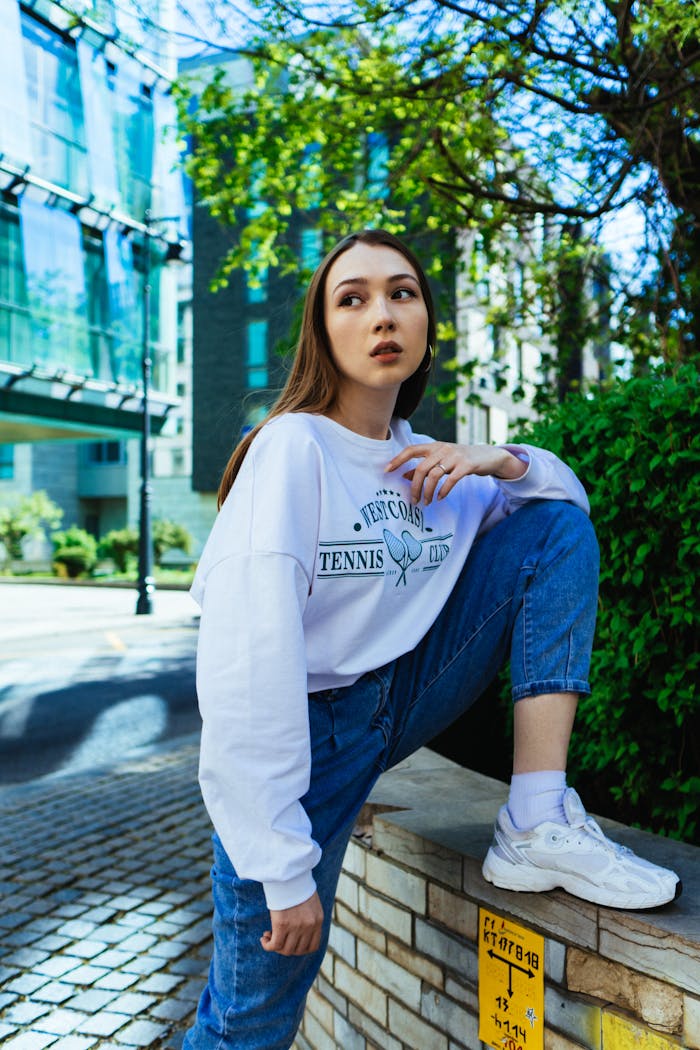 Stylish young woman in denim jeans and sweatshirt outdoors, looking sideways with a fashionable pose.
