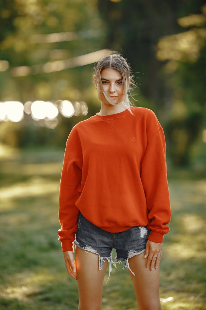 Outdoor portrait of a young woman in a bold orange sweatshirt and denim shorts with natural lighting.